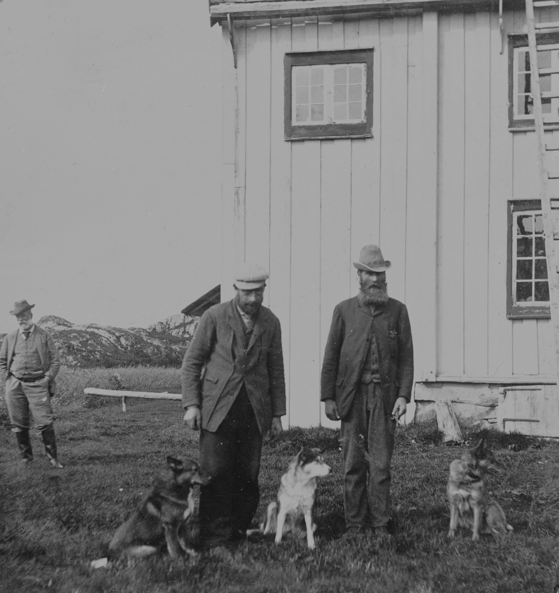 Bildet viser tre menn og tre hunder foran et hus i Svankild, Kvenvær, Hitra. Fra venstre: M.E. Gonne, jaktlos Nils Tungen, og jaktlos Martin Paulsen.