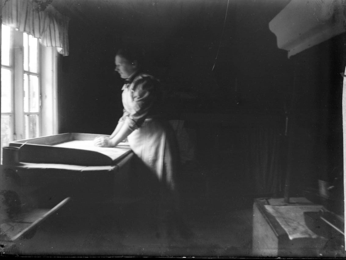 En okänd kvinna står vid ett fönster i ett kök och arbetar på ett bakbord. Bakom henne en spishäll.