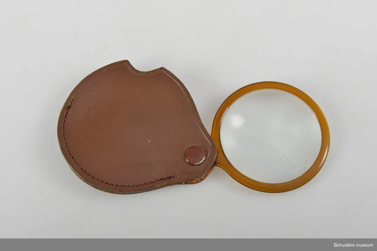 Förstoringsglas. Luppen infattad i brun plast och sitter fästat i ett vridbart brunt läderfodral.