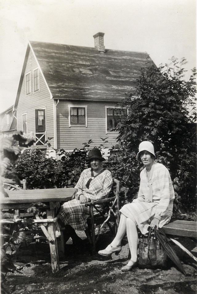 Två kvinnor i hatt och klänning, sitter i en trädgårdsmöbel i en trädgård. 
Bakom dem syns ett bostadshus.