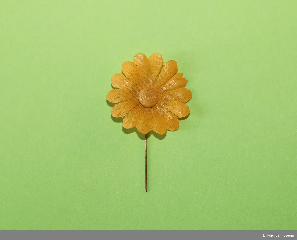 Majblomma från år 1914.
Blomman är gjord av gult, vävt tyg och har 14 blad. Det vävda tyget kallas ”tuskaft”.
I mitten av blomman sitter en gul mittknapp av celluloid. Det som håller blomman samman är en nål av mässing.