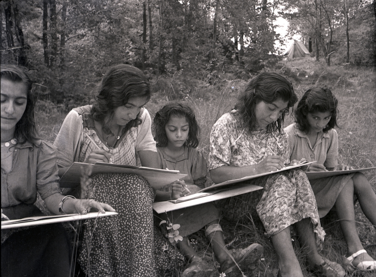 På rad i gräset sitter unga kvinnor och flickor med skrivplattor. I bakgrunden skymtar tält. Bilden är troligtvis tagen i samband med någon av de kurser som arrangerades för romer i mitten av 1900-talet, som kompensation för förlorad skolgång. Stiftelsen Svensk Zigenarmission, grundad 1945, drev sommarskola för romska elever under hela 1940- och 1950-talen. Eleverna var mellan 6 och 60 år och satsningen blev mycket medialt uppmärksammad. Zigenarmissionen var dock kritiserad, både av romer och icke-romer, då man ansåg att romska barn skulle få samma möjligheter att gå i skola som andra svenska barn.  Efter en tid med olika former av försöksverksamhet föreslog den s.k. ”Zigenarutredningen” (1954), att romer skulle gå i vanliga skolor. Frågan om undervisningens organisering fick de enskilda skolorna att hantera på egen hand men från och med juni 1960 fanns möjlighet för kommuner att rekvirera bidrag från staten för speciell undervisning av romska barn. Undervisning för romer arrangerades även av bland annat folkhögskolor och studieförbund. Först efter att alla romer fick bli bofasta på sextiotalet, fick samtliga svenska romer tillgång till skolundervisning.