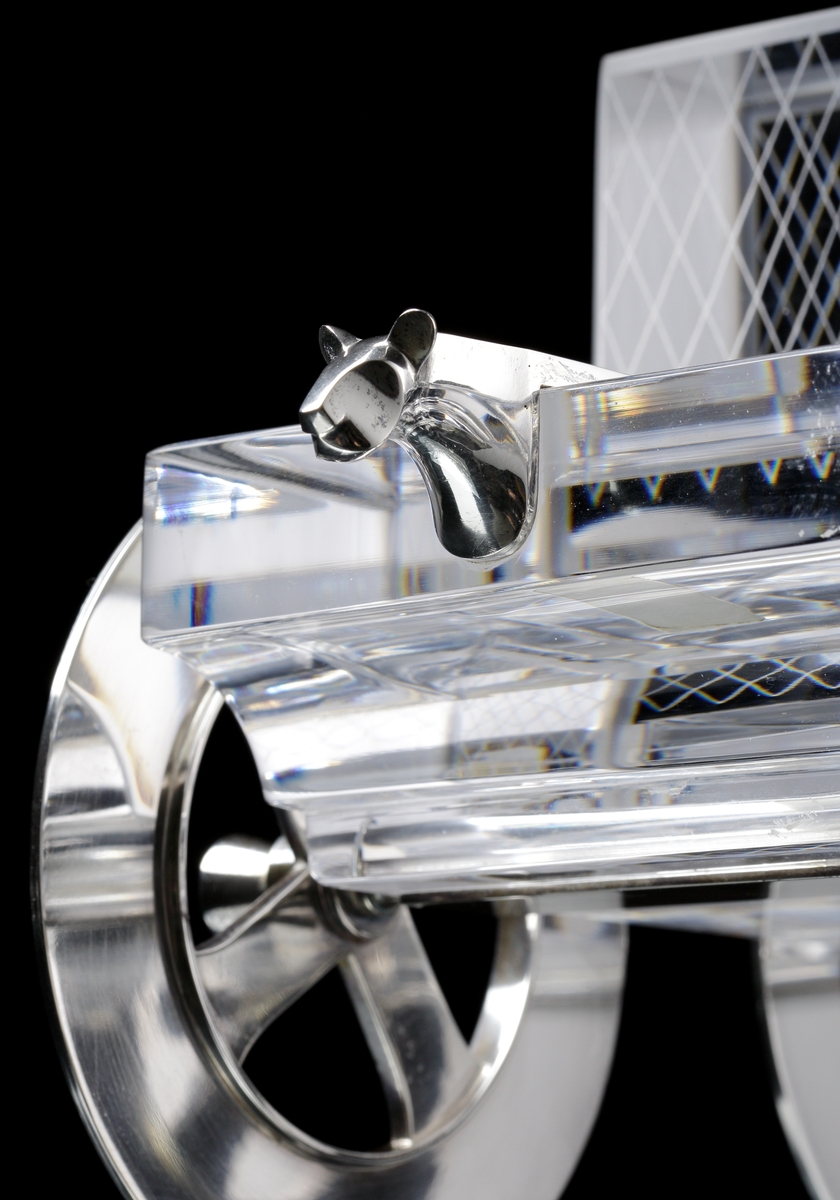 Glasvagn av Gunnar Cyrén, med silverhjul och frontprydnader bestående av jaguarbyster i silver. Karossen är kantig och sittdelen är slipad i ett tunt rutmönster.
