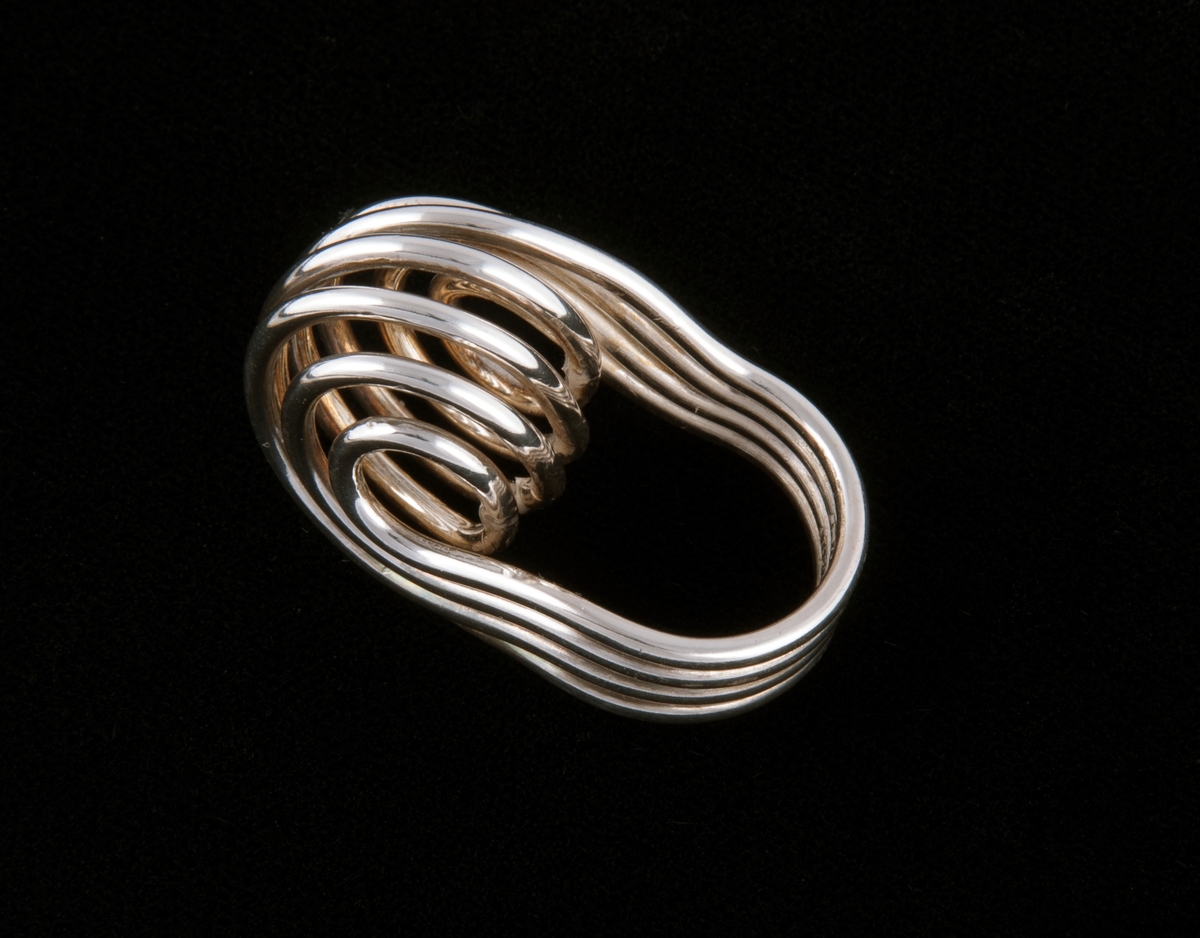 Sammensatt av fire sølvringer som er lagt parallelt til et bånd og vridd i en høy, kuleformet spiral på oversiden.