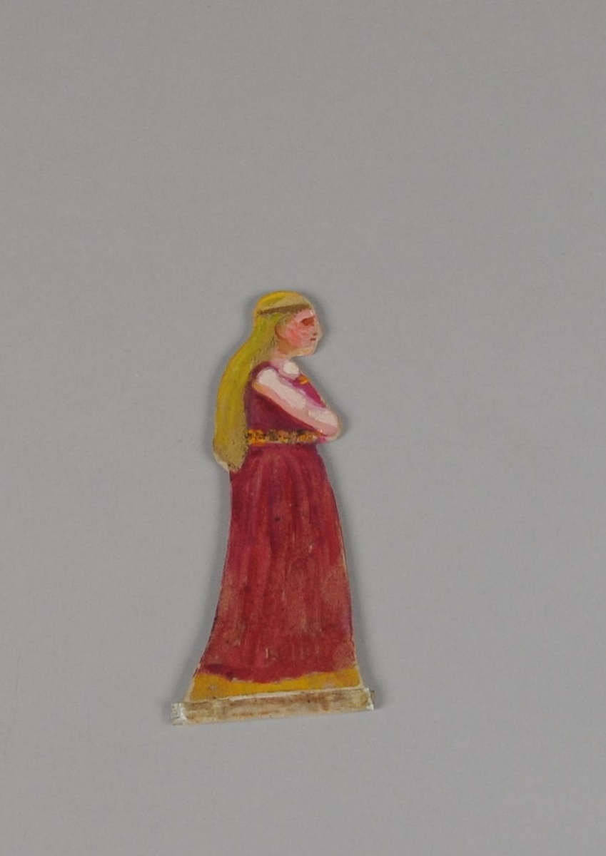 Figur av papp. Forestiller dame i sideprofil, med rød kjole og gullfarget belte. Figuren er malt på begge sider.