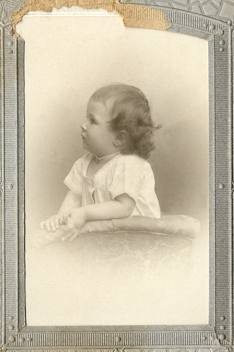 En liten flicka i ljus klänning, med ett sidenband om halsen. Hon vilar armarna mot ryggstödet på en stoppad stol. 
Midjebild, profil. Ateljéfoto.

Fotografens dotter