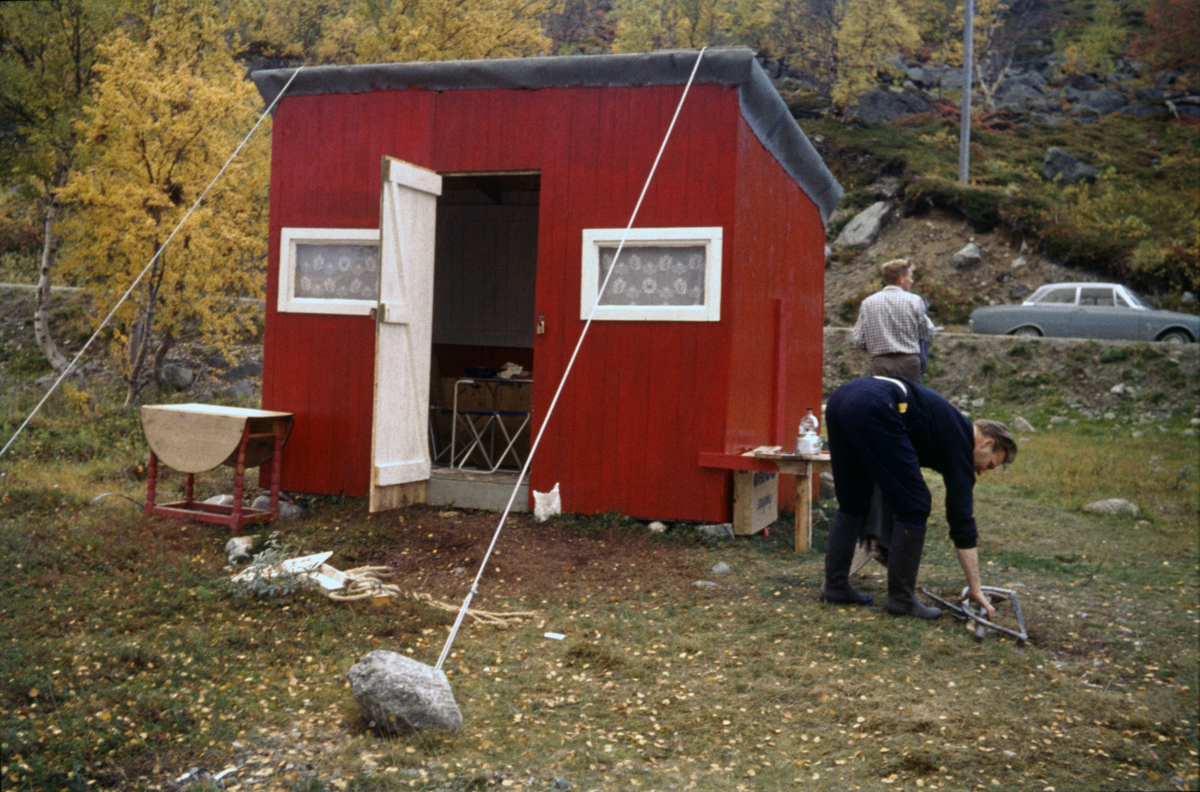 Rødmalt bu med blondegardiner i vinduene. Bua er en del av en enkel sjøflyhavn langs flyruta et sted langs kysten av Troms eller Finnmark. På veien bak bua står flyger Per Lilleengs bil. To av Widerøes ansatte samler sammen verktøy og annet, som etter alt å dømme har blitt brukt til vedlikehold etter endt sesong. Bua har blitt bardunert, malt og fått ny papp på taket.