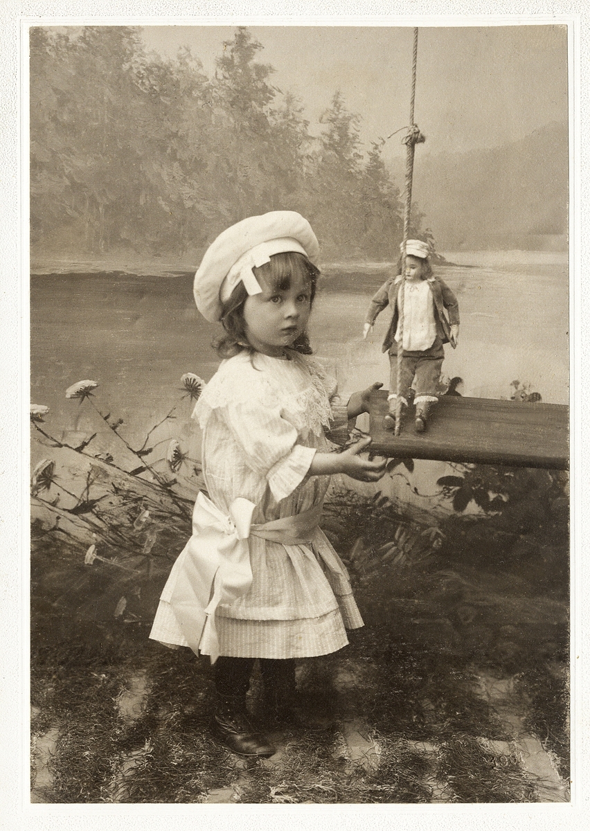 En flicka i ljus klänning och dito mössa. Hon står vid en gunga och leker med en docka.
Helfigur. Ateljéfoto.

Fotografens dotter.