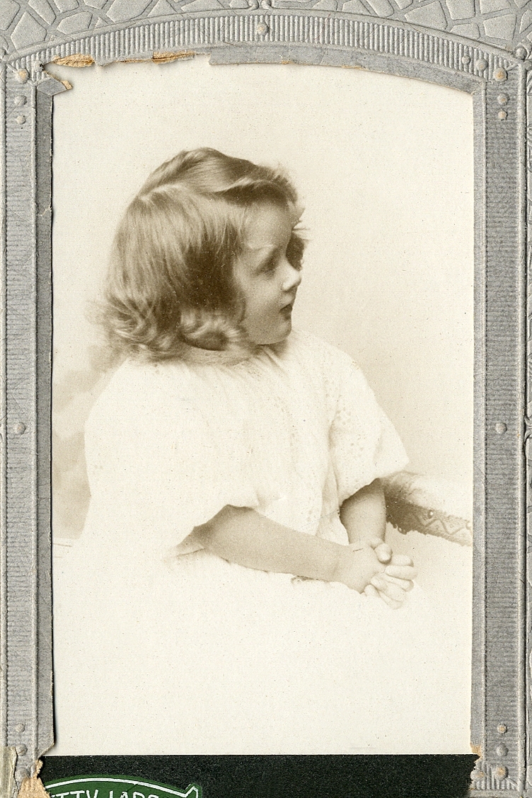En liten flicka i ljus klänning som sitter i en liten soffa.
Knäbild, profil. Ateljéfoto.
Jfr Alb14-091

Fotografens dotter.