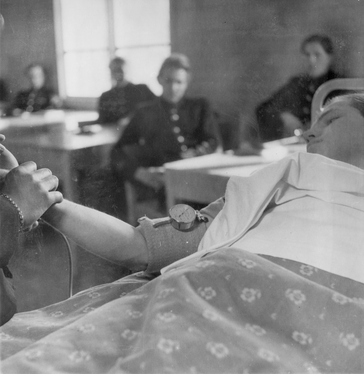 Mätning av blodtryck under sjukvårdskurs på F 11 Södermanlands flygflottilj, 1945. En man i en sjuksäng i skolsal. Elever i skolbänkar i bakgrunden.

Ur fotoalbum "Sjukvårdsskolan 15/1-15/3 1945" från F 11.