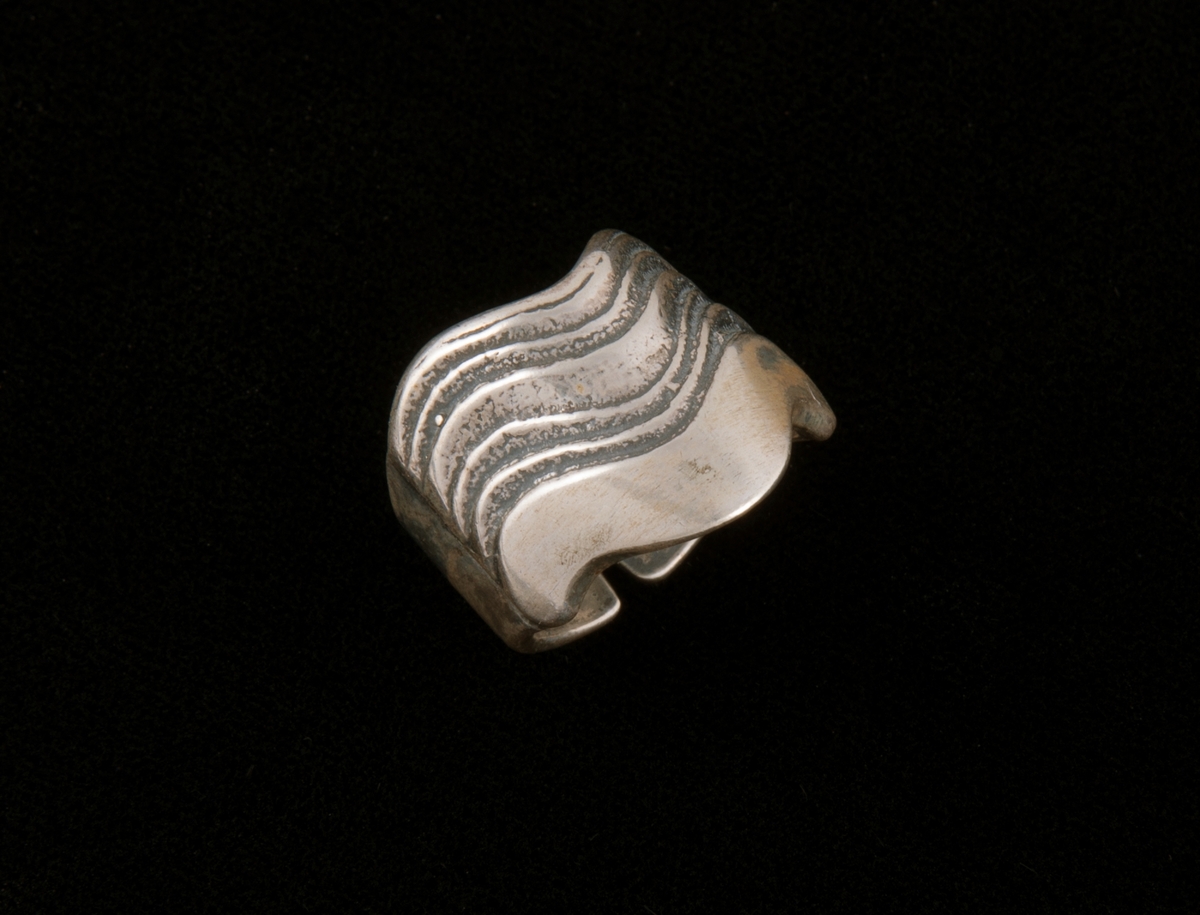 Støpt, blankpolert ring av bredt sølvbånd med regulerbar åpning på undersiden og dekorert parti på oversiden. Dekor av 5 parallelle nedhengende og stiliserte bølger i ringens lengderetning. sidekantene buer på samme måte.