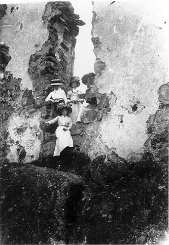 Fyra kvinnor i ljusa klänningar och stora, vida hattar står och sitter i en glugg Brahehus-ruinen.