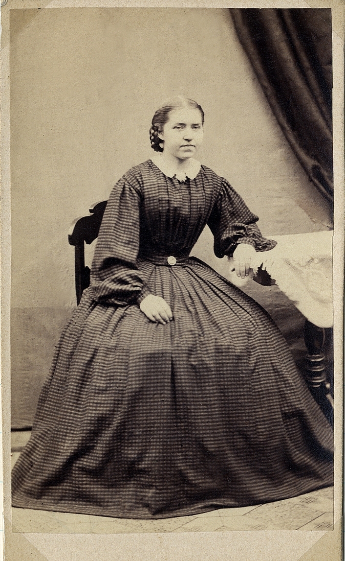 Porträttfoto av en kvinna i mörk, smalrandig klänning (krinolin) med vit krage. Vid kragen syns en brosch. 
Hon sitter på en stol vid ett pelarbord med en spetsduk på. 
Helfigur, halvprofil. Ateljéfoto.
