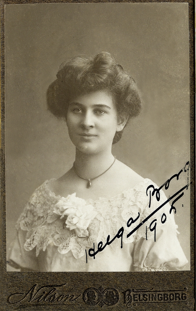 Porträttfoto av en ung kvinna i festklänning med spetskrage m.m. Runt halsen syns en kedja med en berlock. 
Snett över nedre högra delen syns en autograf: "Helga Borg, 1905".