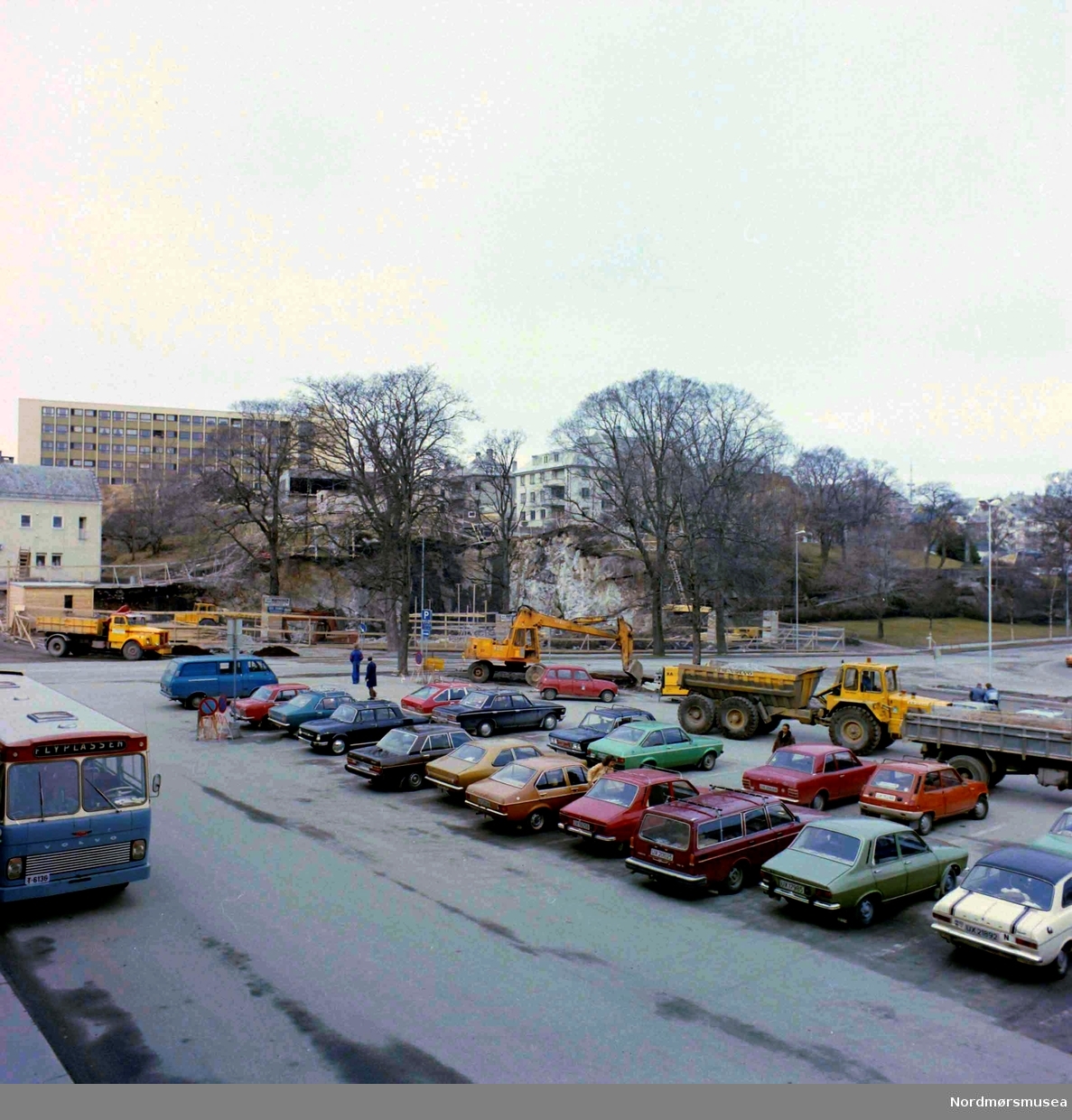 Nerparken 1977. serie.  Bussen til flyplassen avgår fra Braaten SAFE kontor i Tinghuset. Til vestre på bildet ses en buss fra Kristiansund Frei Billag. Selskapets busser var lakkert i grå-blå med rødt tak og gule felt rundt vinduene. Denne bussmodellen ble levert ca. 1970-1974 og viser at bildet ikke er tatt før 1970. (info: 
Sveinung Berild)   -Foto fra parkeringsplassen på Kongens plass på Kirkelandet i Kristiansund, hvor vi i bakgrunnen ser pågående arbeid i det som i dag er Sparebank1 Nordvest sin tomt i Langveien 21.  Fotograf er Nils Williams. Fra Nordmøre museums fotosamlinger.