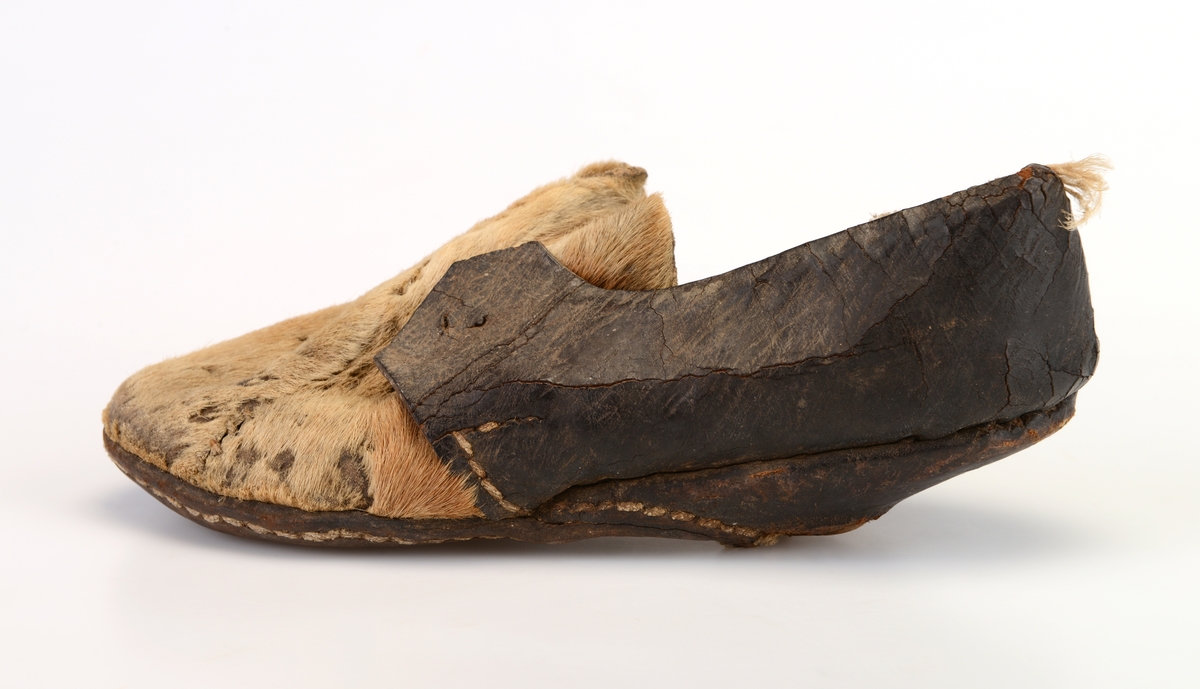 En sko av brunsvart skinn og beige hundeskinn/pels. Den bakre delen er av brunsvart skinn. Den remre delen/tåen er av beige hundeskinn med pelssiden ut. Det er en flik av det brunsvarte skinnet som er brettet over hundepelsen på venstre side av skoen. Innersålen er av brunsvart skinn og skoen er foret med briege strie. Yttersålen er sv brunsvart skinn og den er sydd på med hvit kraftig tråd, Hælen er av tre (knekt).