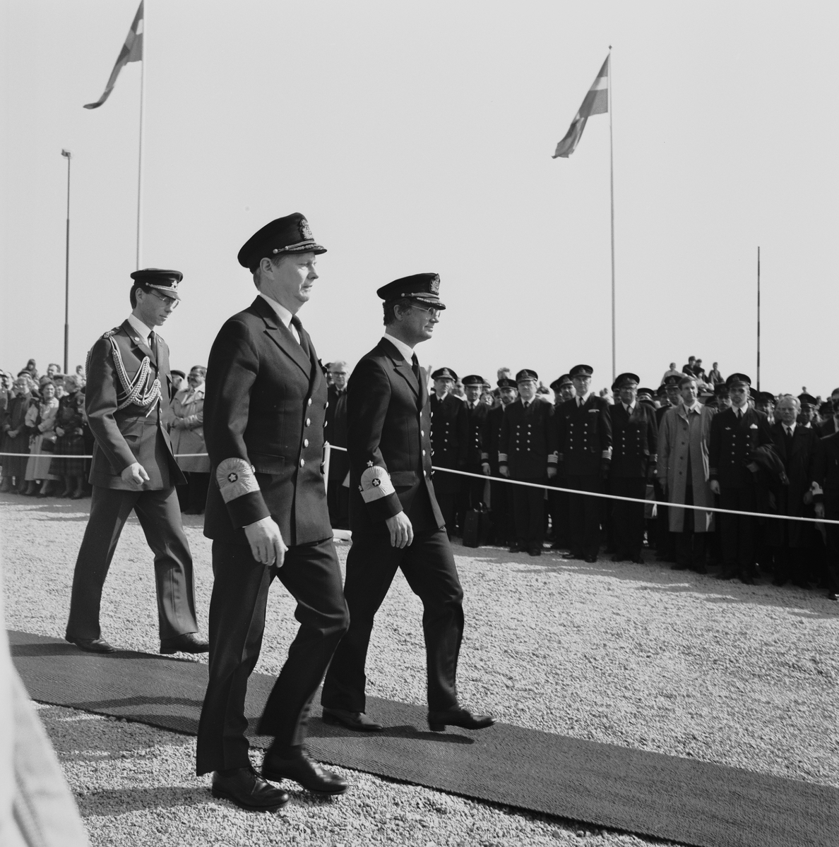 Marinchefen, viceamiral Bengt Schuback och kung Carl XVI Gustaf i amiralsuniform skrider över röda mattan under en sjösättningsceremoni. I bakgrunden syns en åskådarmängd av civila och militärer.