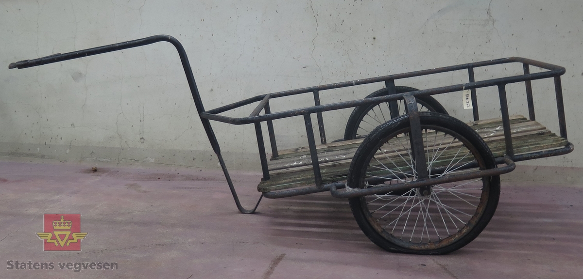 Tohjuls tilhenger for transport etter sykkel, Svart ramme og kontruksjon. Gulv av tre, har rester av grønn farge. Preget av rust.