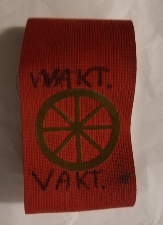 Rødt armbind med olivengrønt vognhjul, og ordet "VAKT" skrevet på for hånd.