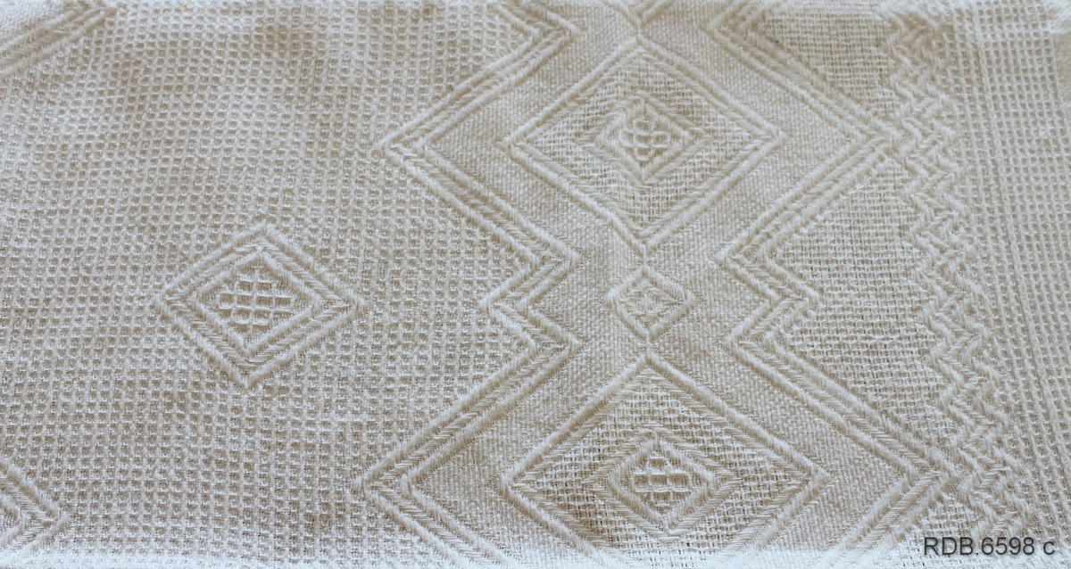 Fabrikkvevd sengeteppe i kvit bomull. Geometriske mønster i vevinga. Påknyttede frynser rundt kanten.