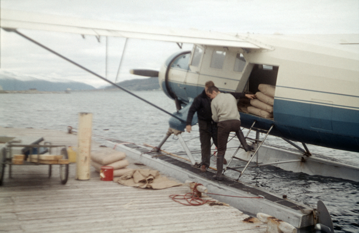 Nor-Wings utfører et transportoppdrag, trolig i forbindelse med vannkraftutbygging på Helgeland. To flygere laster sement ombord i en Norsman som ligger fortøyd til ei flytebrygge ved kysten.