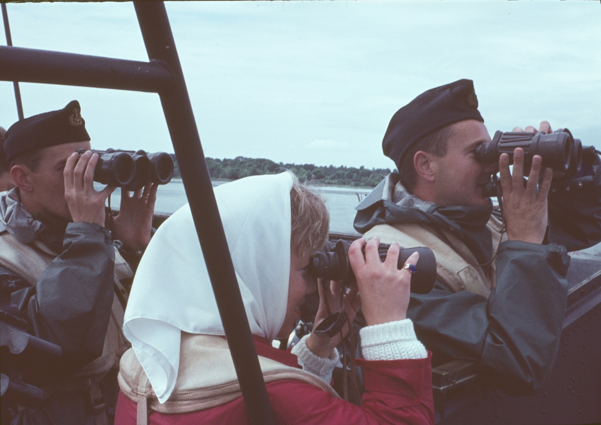 Bilden visar två besättningsmän och en kvinnlig besökare i skyddskläder ombord på en motortorpedbåt. Alla håller utkik med kikare.