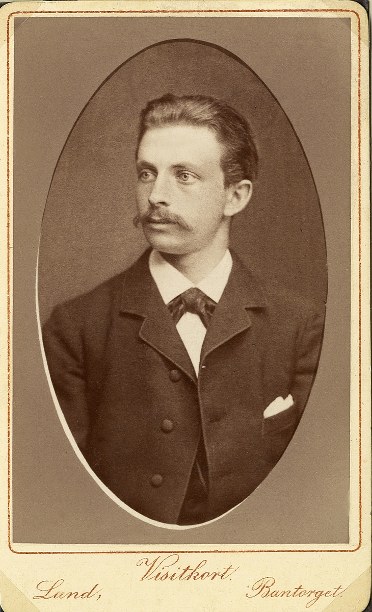Porträttfoto av en ung man med mustascher, klädd i mörk kavajkostym med fluga. 
Bröstbild, halvprofil. Ateljéfoto.