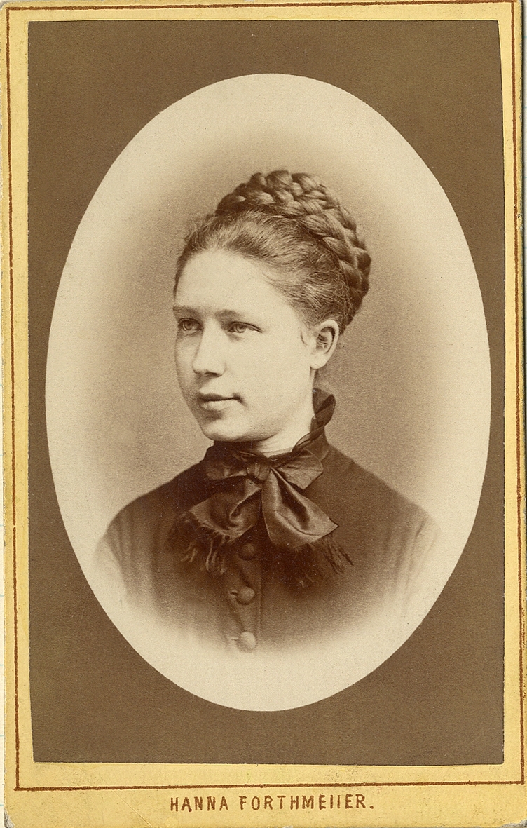 Porträttfoto av en ung kvinna i mörk klänning med en kort mörk sidenscarf runt halsen. 
Bröstbild, halvprofil. Ateljéfoto.