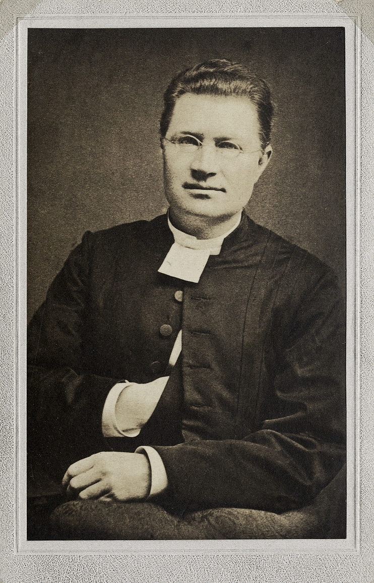 Foto av en man med glasögon, klädd i prästrock och prästkrage.
Midjebild, halvprofil. Ateljéfoto.