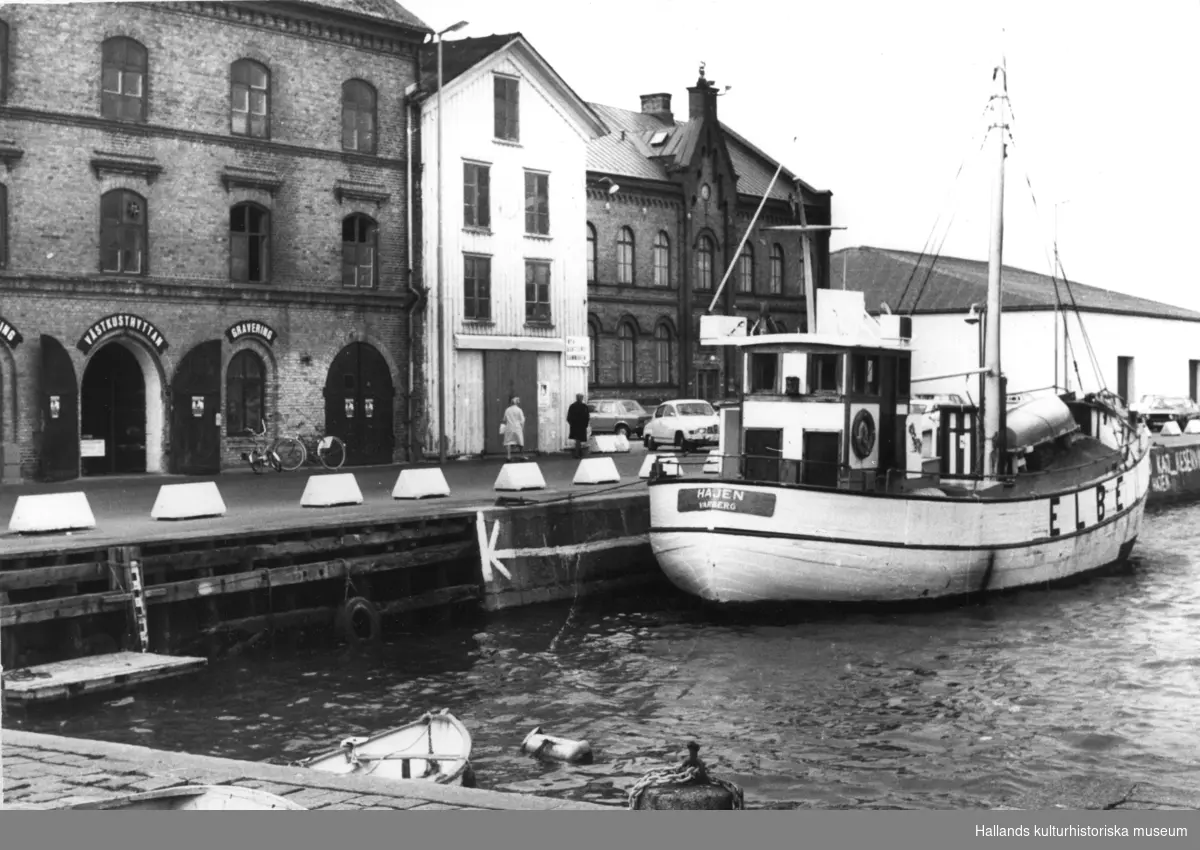 Bild från hamnen, Varberg. Från vänster: byggnad som inrymmer glashytta, gravering, affär med fiskeutrustning och försäljning av fisketurer, auktionslokal m m. Magasinet av trä i bildens mitt revs i april-maj 1978. Längst till höger syns tullhuset och vid kaj ligger båten Hajen från Varberg, märkt ELBE på sidan. Uppgifter saknas om fotograf och när fotot är taget.