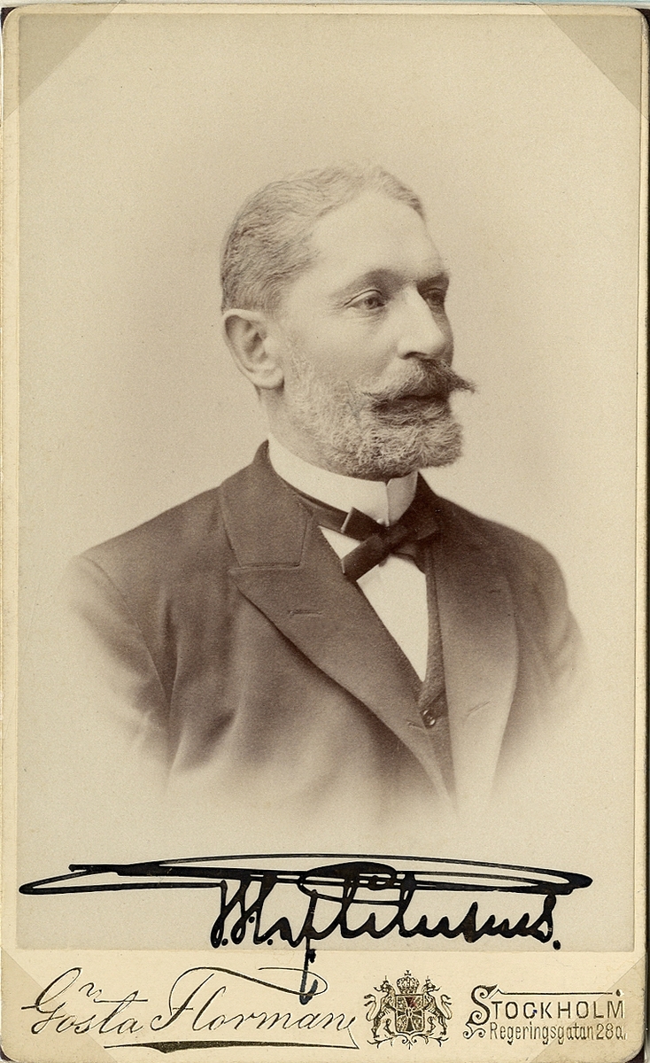 Foto av en medelålders man med mustascher och hakskägg. Han är klädd i mörk kavajkostym med väst, stärkkrage och fluga. 
Under fotot syns en autograf: "G.H. af Petersens".
Bröstbild, halvprofil. Ateljéfoto.