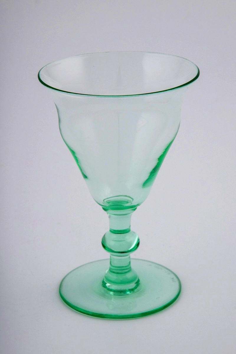 Vinglass av gjennomskinnelig glass med grønnlig tone. Buktende klokkeformet kupa, stett med vulst, svakt skrånende fotplate.