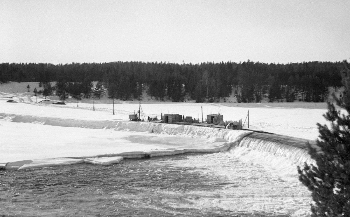 Kraftverksdammen ved Skjefstadfossen i Heradsbygda i Elverum.  Fotografiet er tatt i februar 1942, på et tidspunkt da bare den østre, djupeste og mest vannrike delen av elveleiet var åpent.  Den vestre delen av elveleiet derimot, var is- og snødekt.  Fotografen har stått på Glommas østre bredd med kameraet vendt mot den buete damterskelen.  Om lag midt på dammen er det reist et par brakker, antakelig i samband med anleggsarbeid som ble gjort vinterstid, da det var liten vannføring.  Vi ser også at det er lagt strøm på midlertidig oppsatte stolper ut mot brakkene, antakelig for å få energi til belysning på arbeidsstedet i den mørke vintersesongen med korte dager.  På motsatt side av vassdraget sto barskogen tett. 