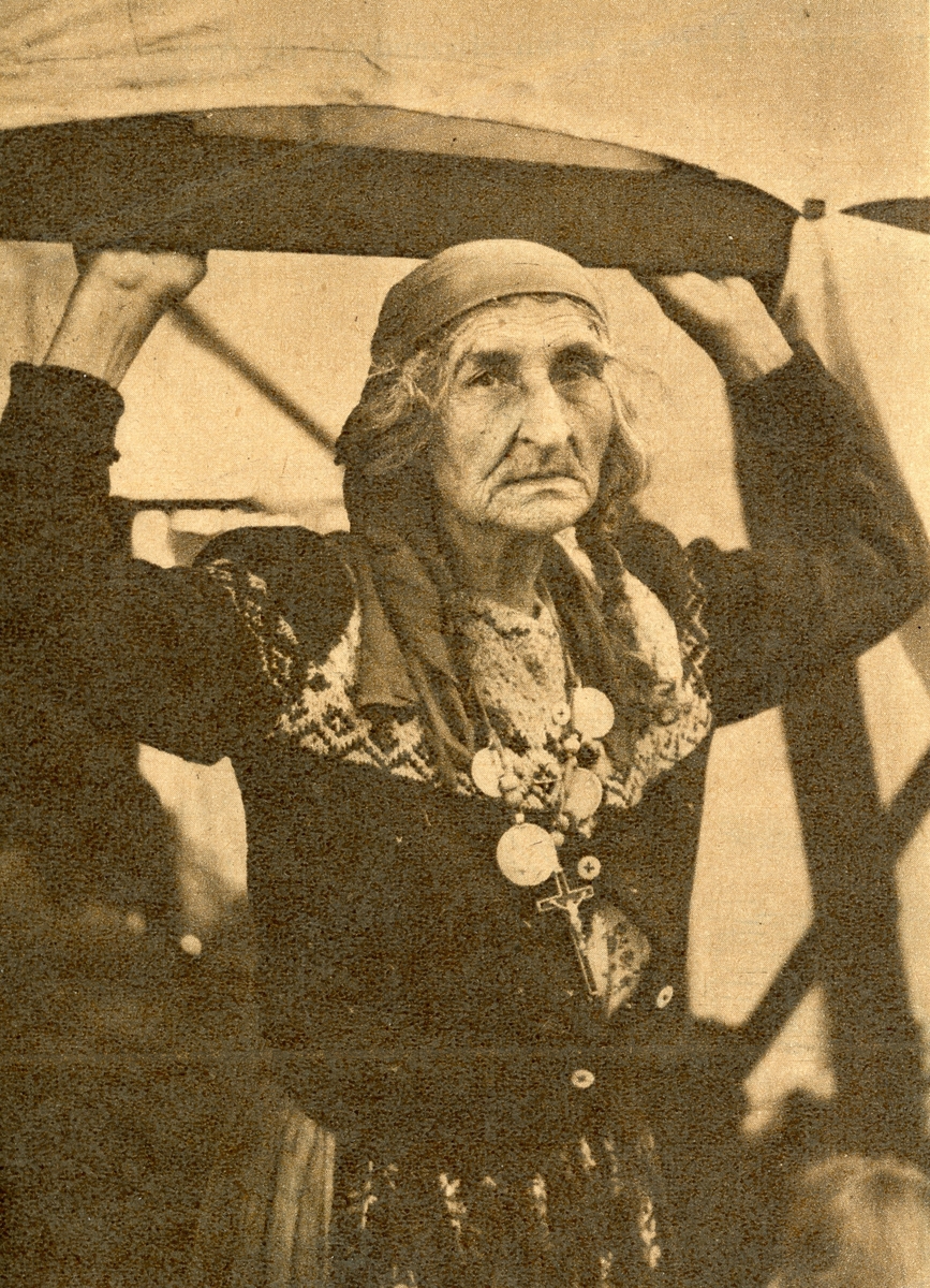 En äldre kvinna med håret i fläta ser in i kameran. Hon ser allvarlig ut och stöder sig mot en tältkonstruktion. Runt hennes hals hänger ett halsband med mynt och ett krucifix.