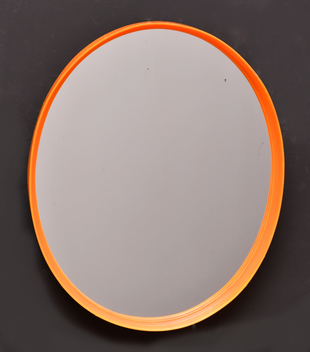 Et ovalt speil. Rammen er av oransje plast. Også opphenget på baksiden av speilet er av oransje plast. Platen bak er av brun plast. Speilet er kjøpt på en DOMUS-butikk for kroner 16,50.-.