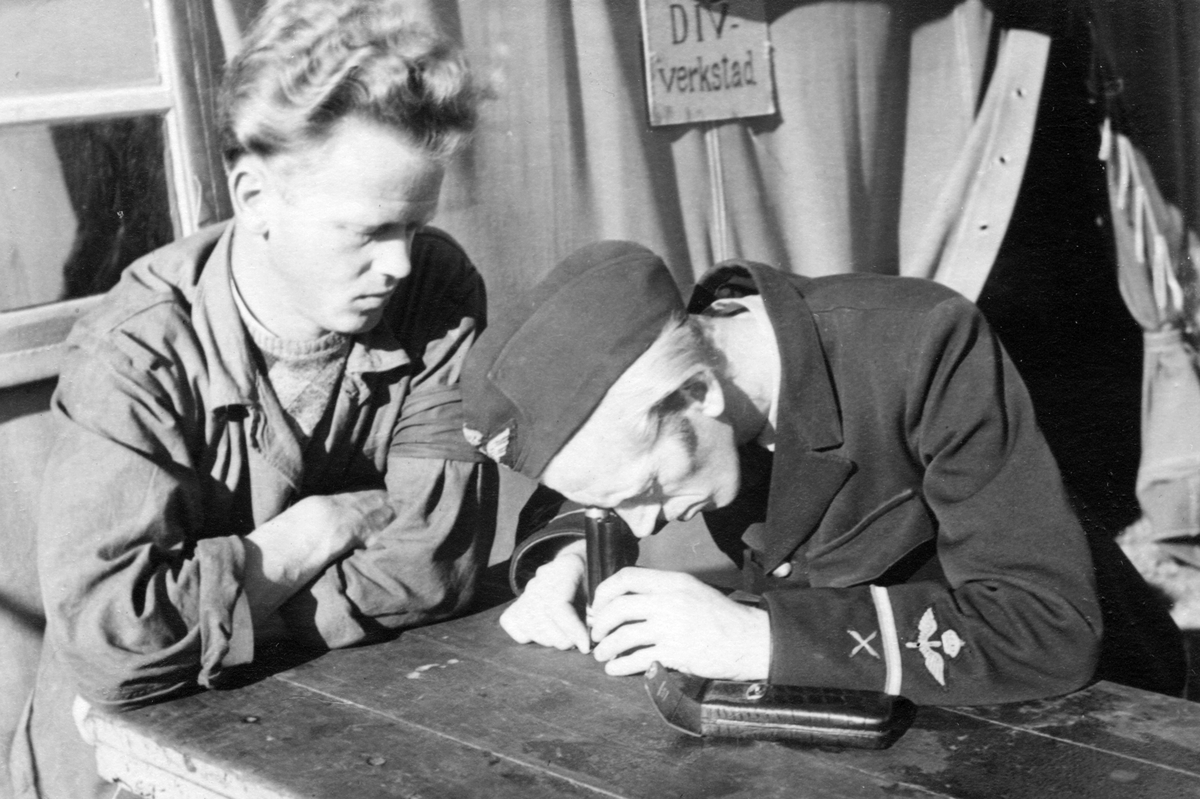 Inspektionsarbete på divisionsverkstad, 1940-tal. En flygmekaniker undersöker ett föremål objekt med förstoringsglas. Bredvid honom sitter en man i overall.