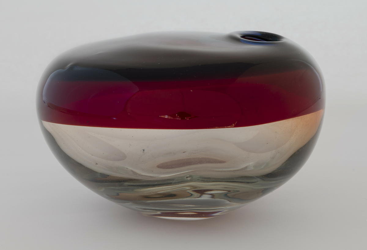 Ovalformet vase i tykt glass. Nedre delen er utført i klart ufarget glass. Den øvre delen er utført i rødfarget opakt glass, som graderes mot blå- og rødtonet klart glass rundt vasens åpning.