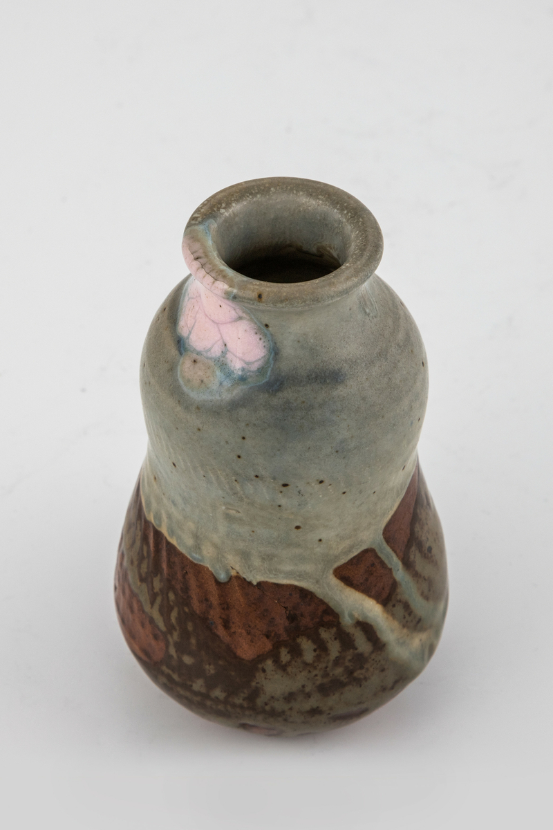 Liten kalebasformet vase i glasert steingods. Den øvre delen er dekket med en blågrå glasur med rosa partier rundt munningsranden. På nedre del renner en gråbeige glasur over en brunrød bunn. Spiralformede riller på nedre del av korpus.