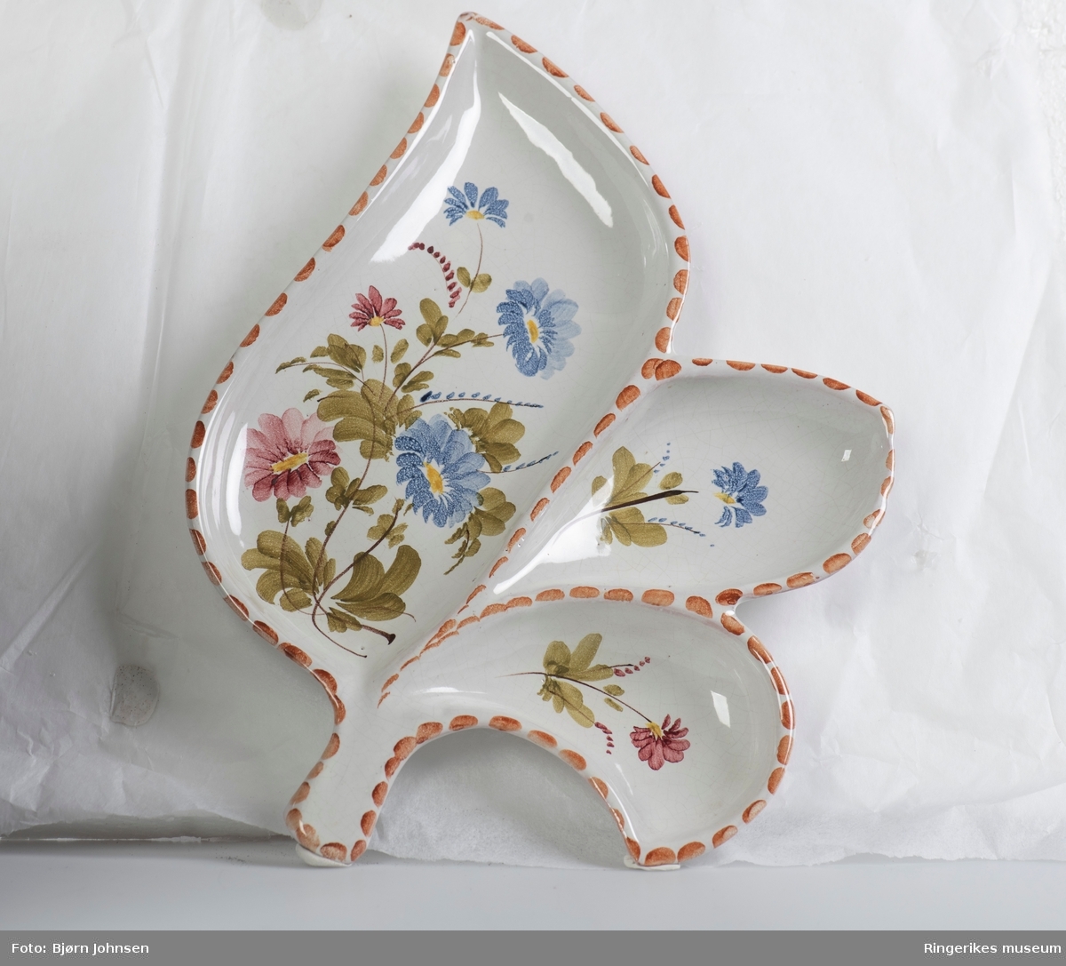 Keramikkfat formet som et blad, med ett stort og to små rom. Dekorert med håndmalte blomster. Stilken på bladet er knekt av og mangler. Antakelig produsert av Arol keramikk i Halden.