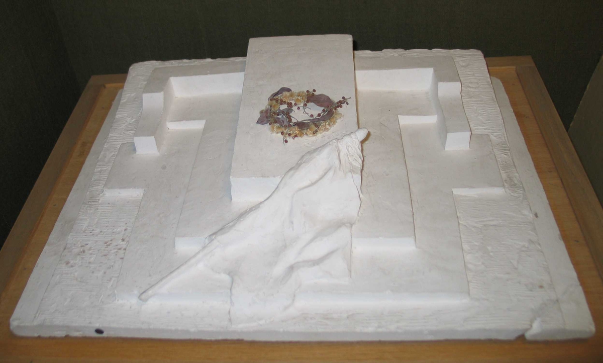 Modell av et gravsted støpt i gips. En gravplate ligger på skrå på et platå med flere avsatser, flankert av murer. Et flagg med sørgeflor ligger dandert over graven.