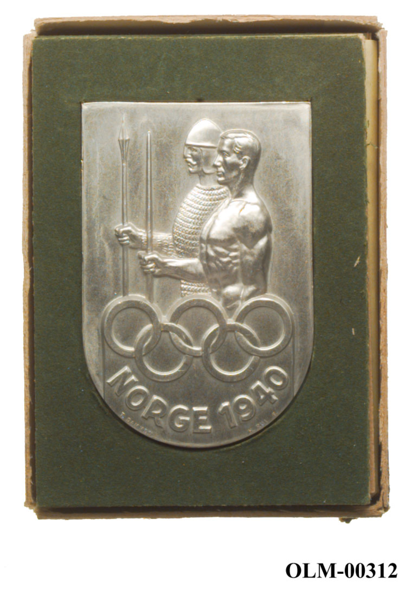 Sølvfarget plakket med motiv av to mannspersoner. Den ene bærer en form for rustning og hjelm. De olympiske ringene inngår også i motivet.