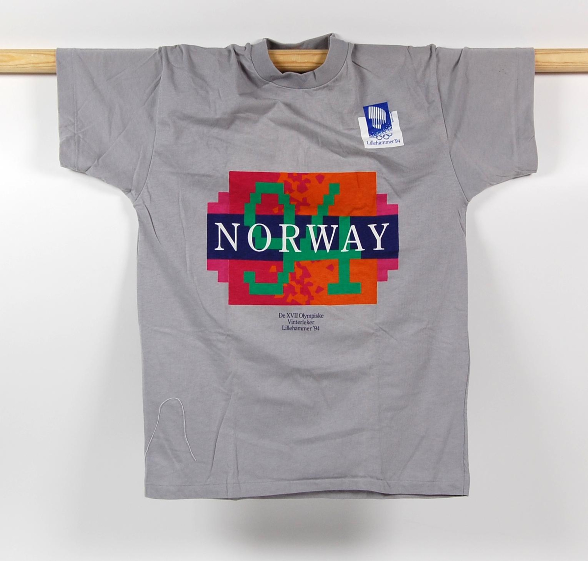 Grå t-skjorte med 2 logoer for de olympiske vinterleker på Lillehammer i 1994. Den ene logoen er flerfarget og har krystallmønster. Krystallmønsteret inngikk i LOOCs designprogram.