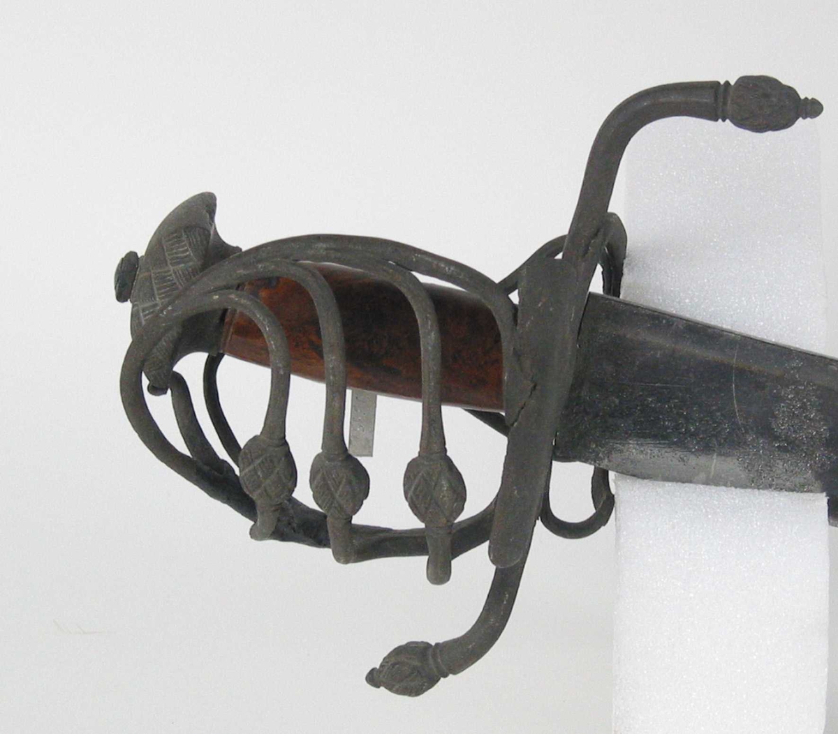 Pyramideformet knapp med kryssfilt dekor og asymmetrisk feste med 3 sidebøyler med fortykninger på midten på utsiden og en på innsiden. Nyreformet parerplate på utsiden og klingebrekker og tommelbøyle på innsiden. S-formede, runde parérstenger med fortykning med filt dekor og dråpeformet avslutning.
Tregrep. Enegget krum, kileformet klinge med en ryggstillt blodrand. Ryggslipt mot spissen. Spiss brukket. Tre hull på spiss. Ett smedstempel.

