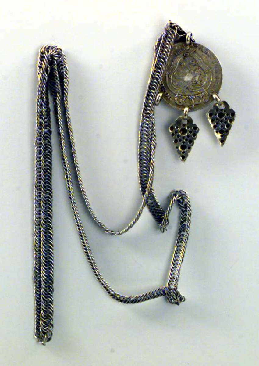Halskjede i metall med sølvmynt som anheng. Fra mynten henger to løv, et tredje mangler. Mynten har krone og kongemonogram.
