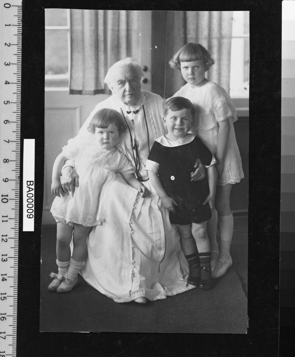 Gruppefotografi; En gammel dame sittende i hvit lang kjole med tre barn stående rundt seg; en gutt i mørke korte bukser og bluse med hvit krave; de to pikene kledd helt i hvitt, i tynne helt korte kjoler med fint broderi. 