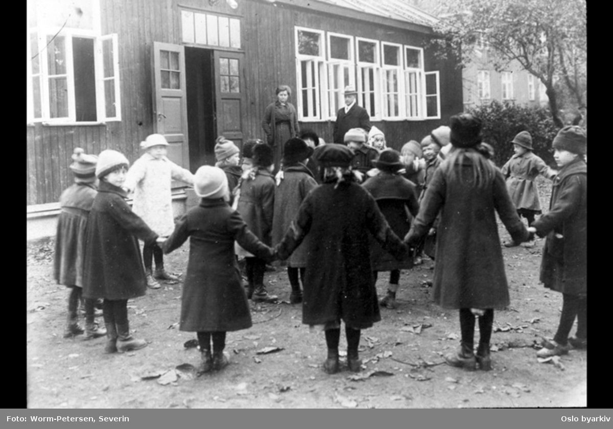 Sandaker friluftsskole. Barn som leker foran skolebygningen. I bakgrunnen Carl Schiøtz og skolens bestyrer Helga Kjerulf.