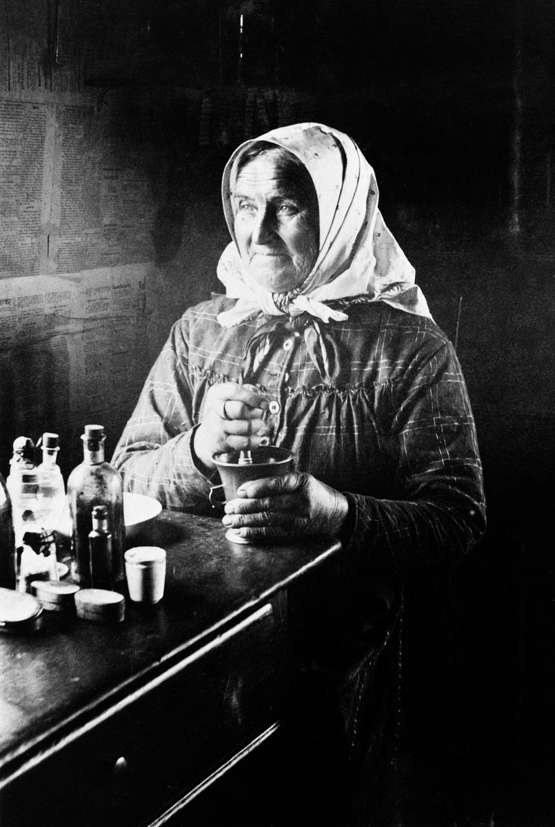 Portrett av eldre dame med skaut som sitter med morter ved bord. Kansje hun blander medisin, urter. Veggene er kledd med avispapir.
