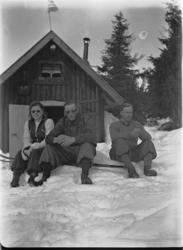 Ukjent kvinne og to menn utenfor ei HYTTE i skogen.