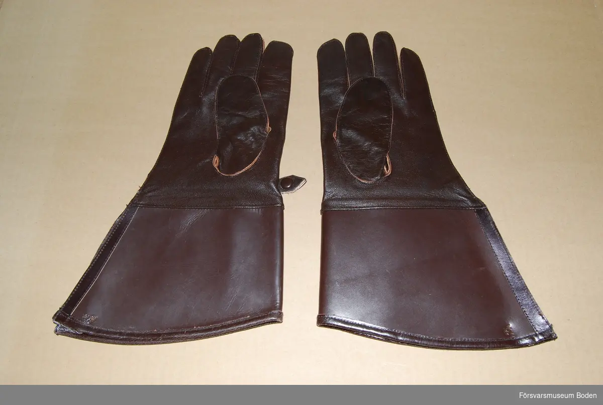 Ett par oanvända handskar av mörkbrunt läder med styv krage som är präglad med tre kronor vid kanten på utsidan. I övrigt ingen märkning. Spänntamp med tryckknapp på ovansidan. Avsedd att användas av bl.a. mc-ordonnanser.