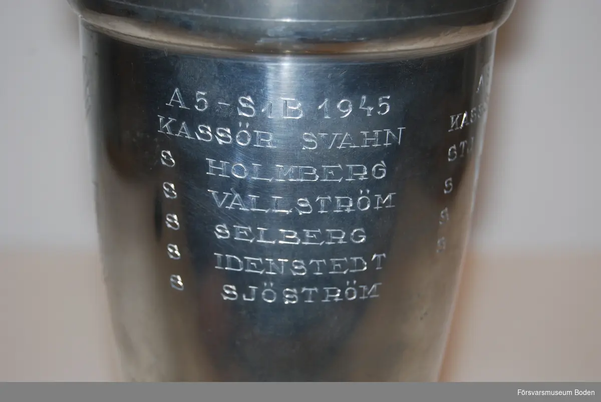 Pokal av tenn på fot av svartlackerat trä. Med avtagbart lock. Pokalen har tre lags inskriptioner: A 5 /S 1B 1945, A 5 1947 och A 5 1948.